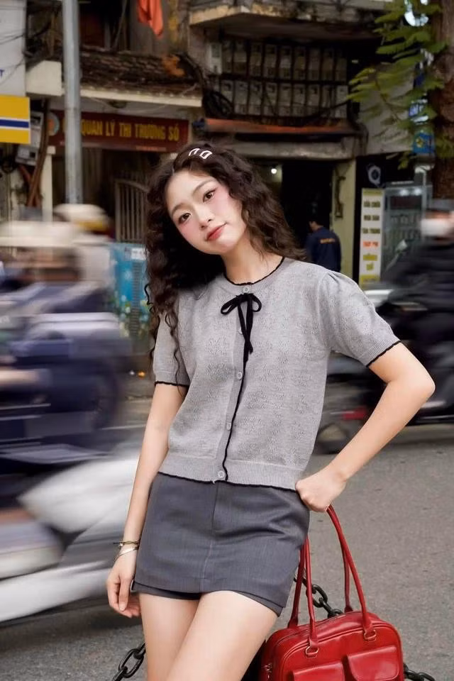 Bộ trang phục là sự kết hợp tinh giản giữa áo len cardigan tay ngắn tông xám trung tính, điểm nhấn là chiếc nơ nhỏ cột cổ màu đen - vừa dịu dàng vừa mang nét tiểu thư hiện đại. Phối cùng chân váy ngắn và túi xách đỏ đậm nổi bật, gợi cảm giác thanh lịch nhưng vẫn đầy cá tính. ẢNH: UNI BY HEART