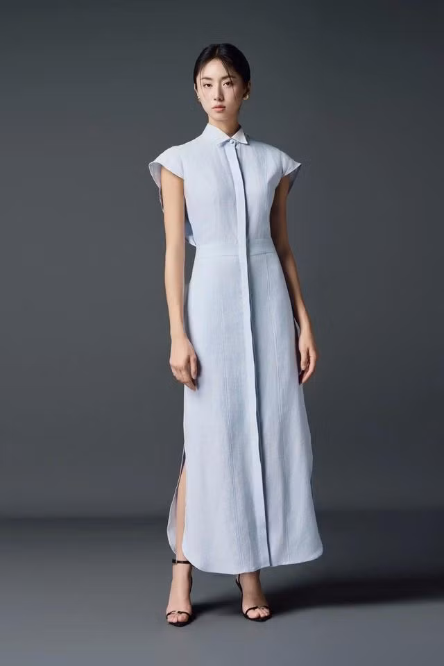 Váy sơ mi dáng dài lấy cảm hứng từ phom dáng của chiếc áo sơ mi không tay tông màu xanh nhạt duyên dáng. Chi tiết nẹp cúc chạy dài từ cổ xuống chân váy tạo nét sắc sảo, kết hợp đường xẻ bên hông khơi gợi trí tò mò của người nhìn ngắm. ẢNH: LYP