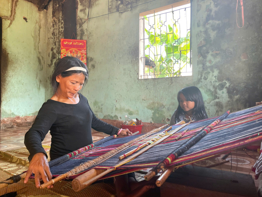 Cô bé Hà My nép mình bên khung cửi, chăm chú nhìn mẹ dệt. Ảnh: Bích Ngọc