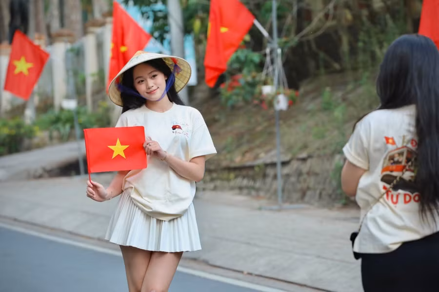 Bạn trẻ khoác lên mình chiếc áo với dòng chữ Độc Lập - Tự Do - Hạnh Phúc và đội nón lá truyền thống check-in con đường cờ hoa.