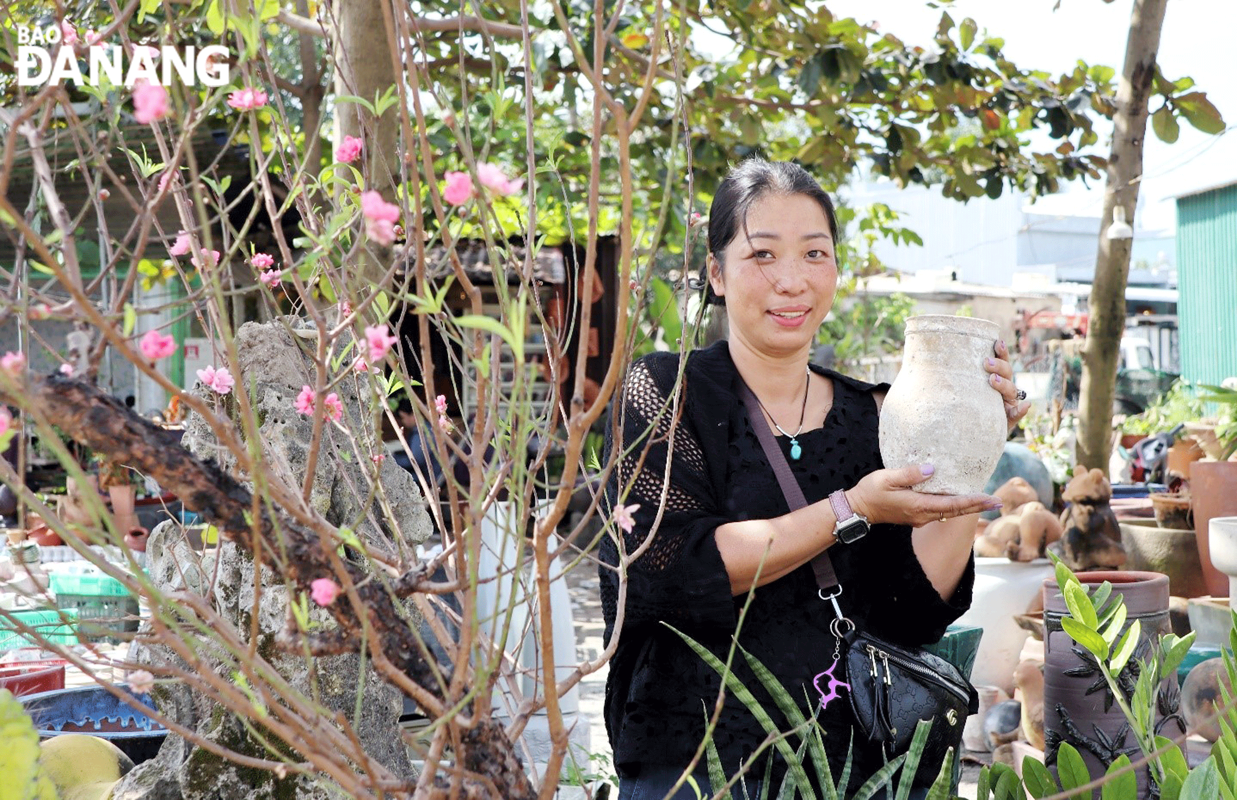 Cô chủ Gốm Nhà Tím giới thiệu một loại gốm giả cổ bên cành đào khoe sắc sớm. Ảnh: NHƯ HẠNH Cô chủ Gốm Nhà Tím giới thiệu một loại gốm giả cổ bên cành đào khoe sắc sớm. Ảnh: NHƯ HẠNH