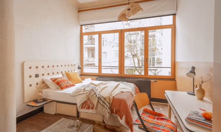 Phòng ngủ 16m2 được thiết kế với tông màu be có cửa sổ lớn đón sáng kết hợp với nội thất đỏ, cam. (Ảnh: Cohabs)