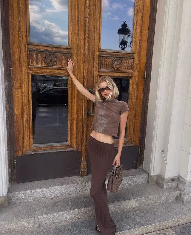 Nữ siêu mẫu gốc Thụy Điển chọn outfit nâu từ trên xuống dưới bằng bản phối chân váy maxi với áo thun crop top khoe vòng eo con kiến. Sử dụng phụ kiện túi xách, kính mắt tiệp màu trang phục là cách mà cô hoàn thiện tổng thể