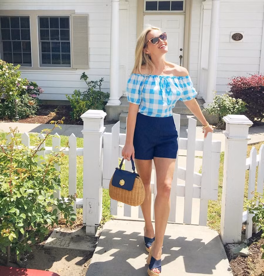  Reese Witherspoon chọn phong cách đơn giản, thoải mái với áo mi trễ vai kẻ ô và quần short cạp cao kết hợp cùng túi xách cói đan kiểu cách.