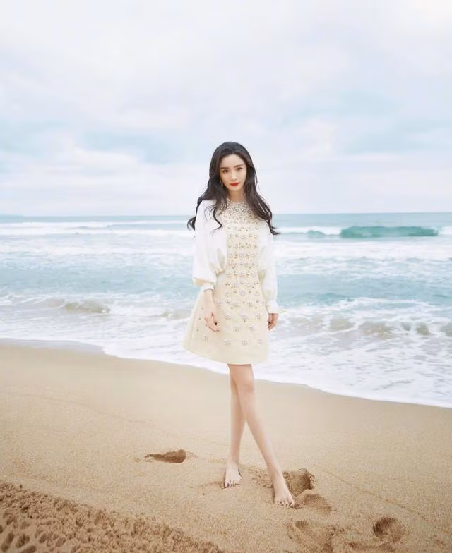 Người đẹp diện váy ngắn màu kem đính ngọc trai để chân trần trên cát, trang điểm tự nhiên với mái tóc xoăn nhẹ bồng bềnh rất nữ tính và ngọt ngào.