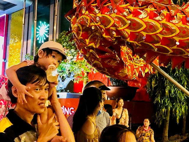 Được bố điệu trên vai, bé gái vừa bú sữa vừa dõi mắt về hướng đoàn Lân đang biểu diễn