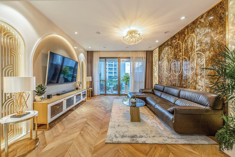 Phòng khách được xử lý với các khung vòm hắt đèn bắt mắt, đối diện là vách đá marble, đan xem hoạ tiết chỉ inox vàng mờ tạo điểm nhấn. Đá marble tự nhiên của Ý còn được sử dụng phối hợp với gỗ tự nhiên để lát cho sàn nhà. Theo đó ở không gian chung liên hoàn là phòng khách – bếp ăn thì phần phòng khách lát gỗ, còn khu bếp ăn lát đá.
