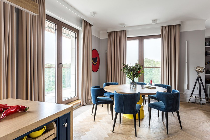 Phía sau phòng khách là bàn ăn. Những chiếc ghế ăn màu xanh bao quanh chiếc bàn ăn bằng gỗ.