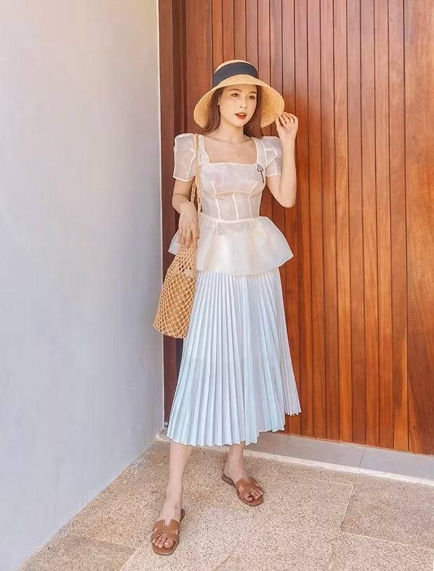 Sam yểu điệu với áo peplum tay phồng và chân váy xếp ly, phối màu sáng ăn ý. Sandals đế bệt, mũ cói vành tròn và giỏ lưới là bộ ba phụ kiện giúp cô nàng hoàn thiện trang phục đi nghỉ.