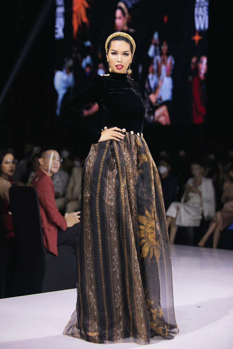 Với vị trí first face mở màn cho buổi diễn, Hà Anh đã tỏa sáng với năng lượng và vẻ quyến rũ hết sức nữ tính, trong đầm dạ hội tối giản với phần thân trên bằng chất liệu nhung the và đầm xòe đính khuy vàng dập nổi logo của thương hiệu. Phần tùng váy được in họa tiết mặt trời tỏa sáng tinh xảo trên nền vải tơ dệt với sợi kim tuyến vàng.