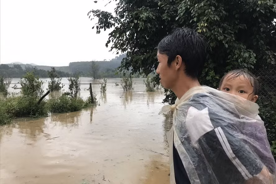 Một người dân xã Cư San, cõng con sau lưng, trùm áo mưa đứng lặng nhìn ra phía lòng hồ Krông Pách thượng - nơi nước lũ đang dâng cao sau bão số 12. Ảnh: Bảo Trung