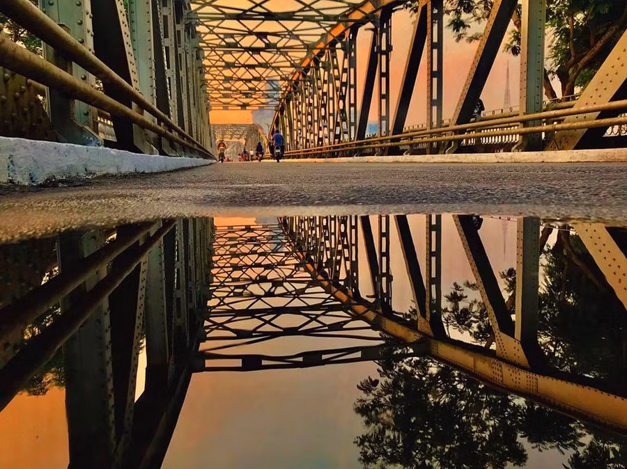 Cầu Trường Tiền là địa danh không thể bỏ lỡ khi đến Huế. Sau cơn mưa đêm đầu hạ, mặt đường vẫn còn ẩm ướt, công trình phản chiếu lên bóng nước, tạo ra hiệu ứng thị giác độc đáo. Nếu quan sát kĩ, bạn sẽ nhận ra những thanh cầu đổi màu liên tục từ đậm đến nhạt dần theo từng 