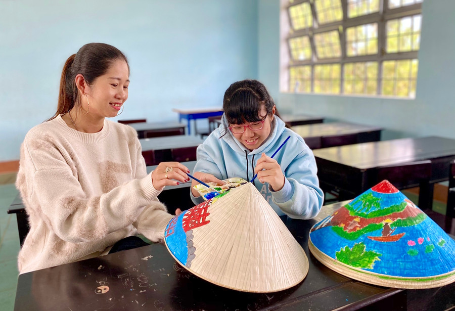 Em Đoàn Huỳnh Thanh Tâm (bìa phải) được cô giáo Mỹ thuật hướng dẫn vẽ trang trí trên nón lá. Ảnh: Mộc Trà