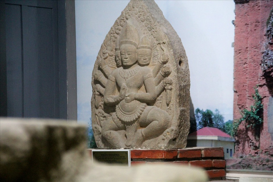 Phù điêu thể hiện thần Brahma trong tư thế nhìn về phía trước, hai chân chùng xuống, hai đầu gối bành ra hai bên, hai tay chính bắt quyết trước ngực. Hai bắp tay, mỗi bên mọc ra thêm ba tay phụ cầm các vật: dao găm, hoa sen. Thần có ba đầu: đầu chính giữa nhìn thẳng, hai đầu hai bên như cố nhô ra để nhìn ra phía trước, cả ba khuôn mặt đều vuông vức, tỏ ra nghiêm nghị.