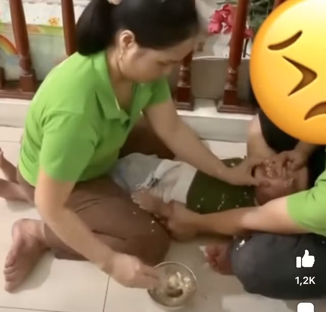Hai bảo mẫu đè em nhỏ ra đất để cho ăn. Ảnh: Cắt từ clip