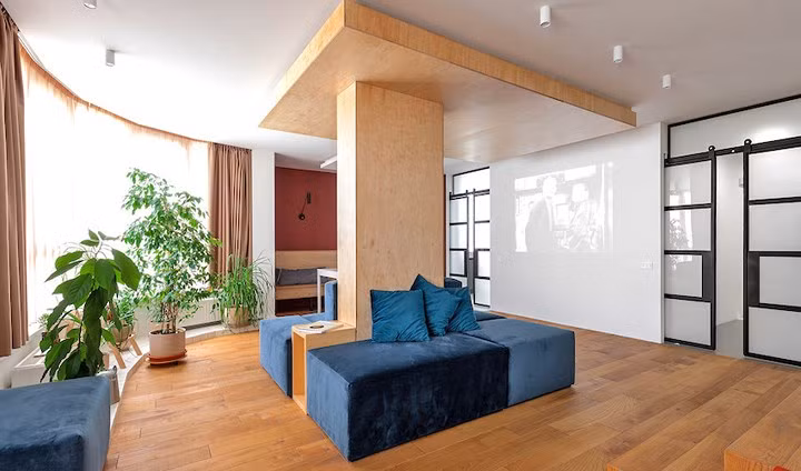 Phòng khách rộng rãi chọn bộ ghế sofa bằng nhung ôm theo trụ đỡ của ngôi nhà, máy chiếu đặt đối diện bức tường màu trắng, cây xanh bố trí men theo khung cửa sổ để lấy ánh sáng và khí trời.