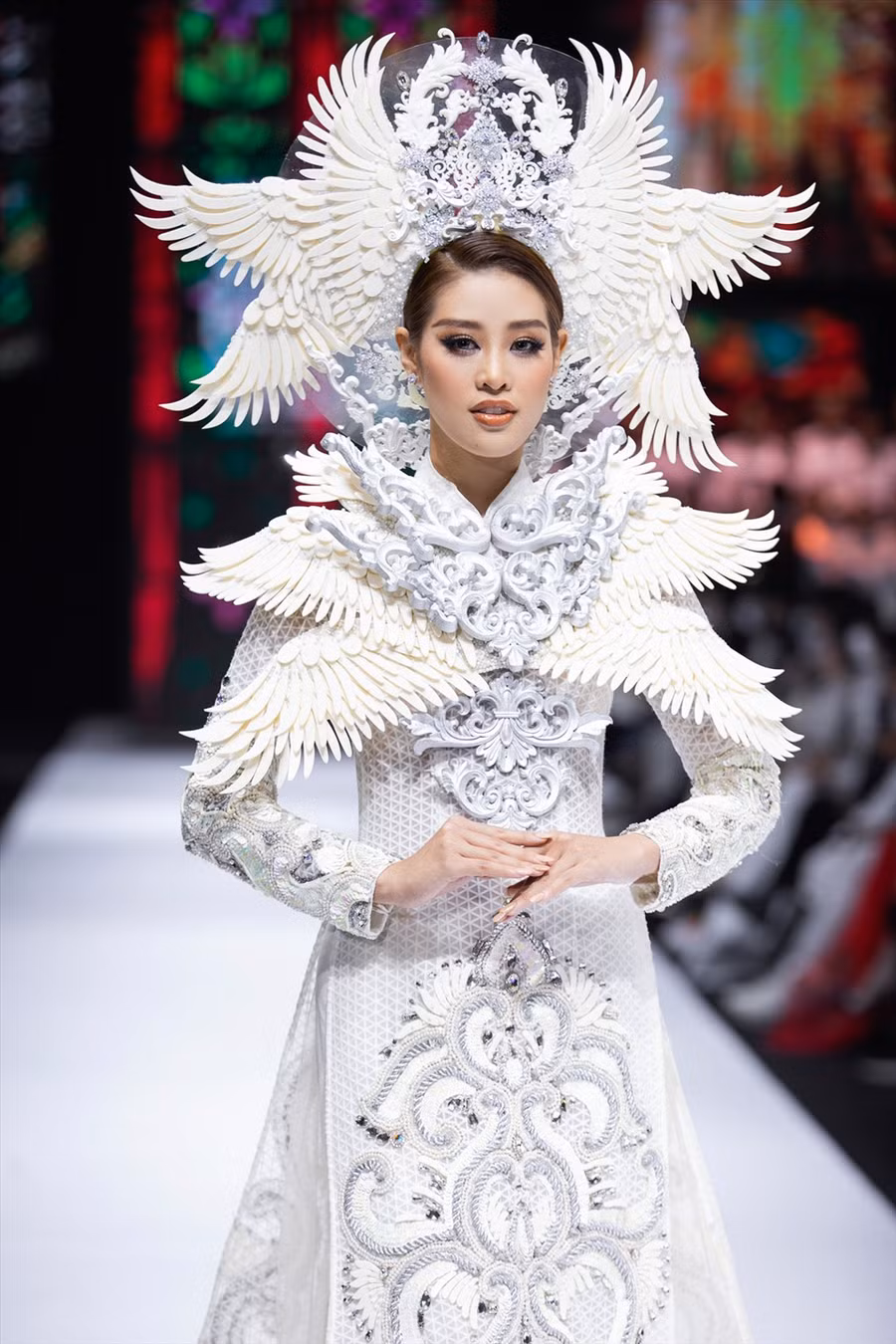 Hoa hậu Khánh Vân chính là vedette của bộ sưu tập Kim Lang. Trên nền nhạc Hallelujah du dương, người đẹp xuất hiện trong tà áo dài cách tân trắng tinh khôi, vạt trước được thêu hoa văn kỳ công cùng chiếc mũ đội đầu là những cánh chim.