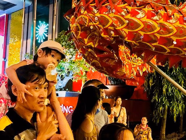 Được bố điệu trên vai, bé gái vừa bú sữa vừa dõi mắt về hướng đoàn Lân đang biểu diễn